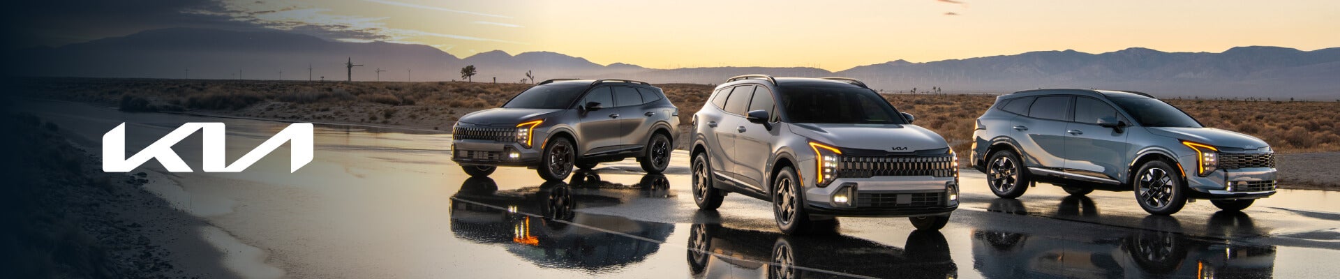 Front three-quarter view of Kia Sportage lineup on wet asphalt with sunset mountains.