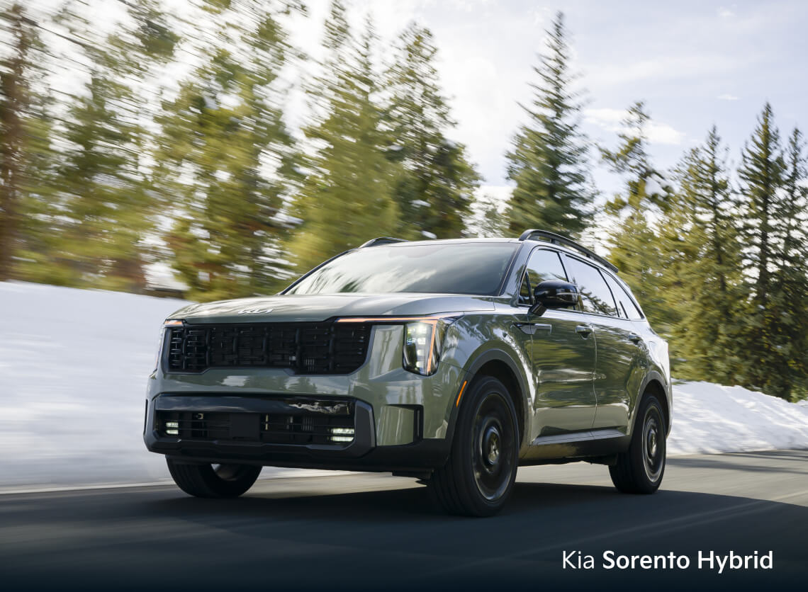Front three-quarter view of sage green Kia Sorento Hybrid EX driving on a snowy mountain road with pine trees.