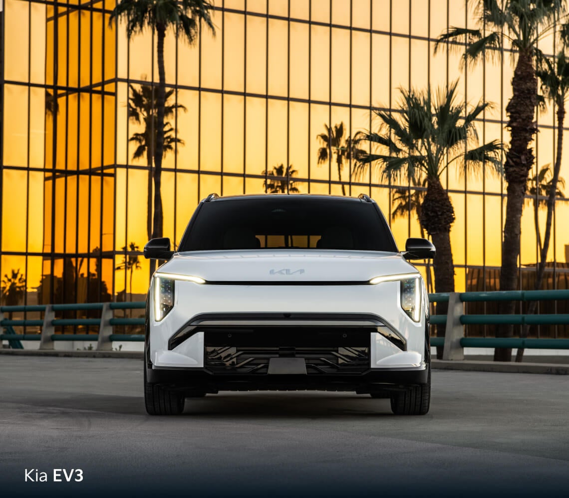 Front view of white Kia EV3 GT-Line parked in front of a glass building with palm trees at sunset.