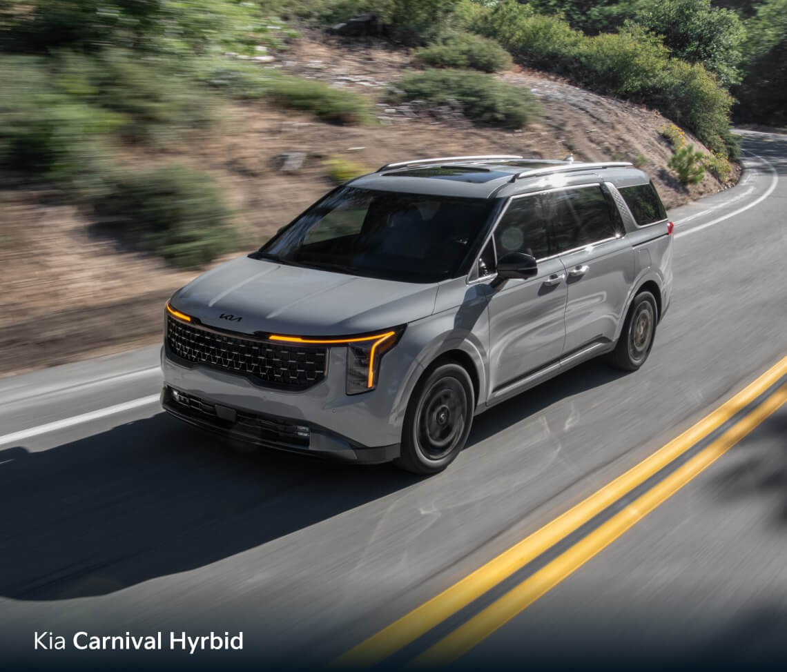 High-angle front three-quarter view of gray Kia Carnival Hybrid driving on a winding forest road.